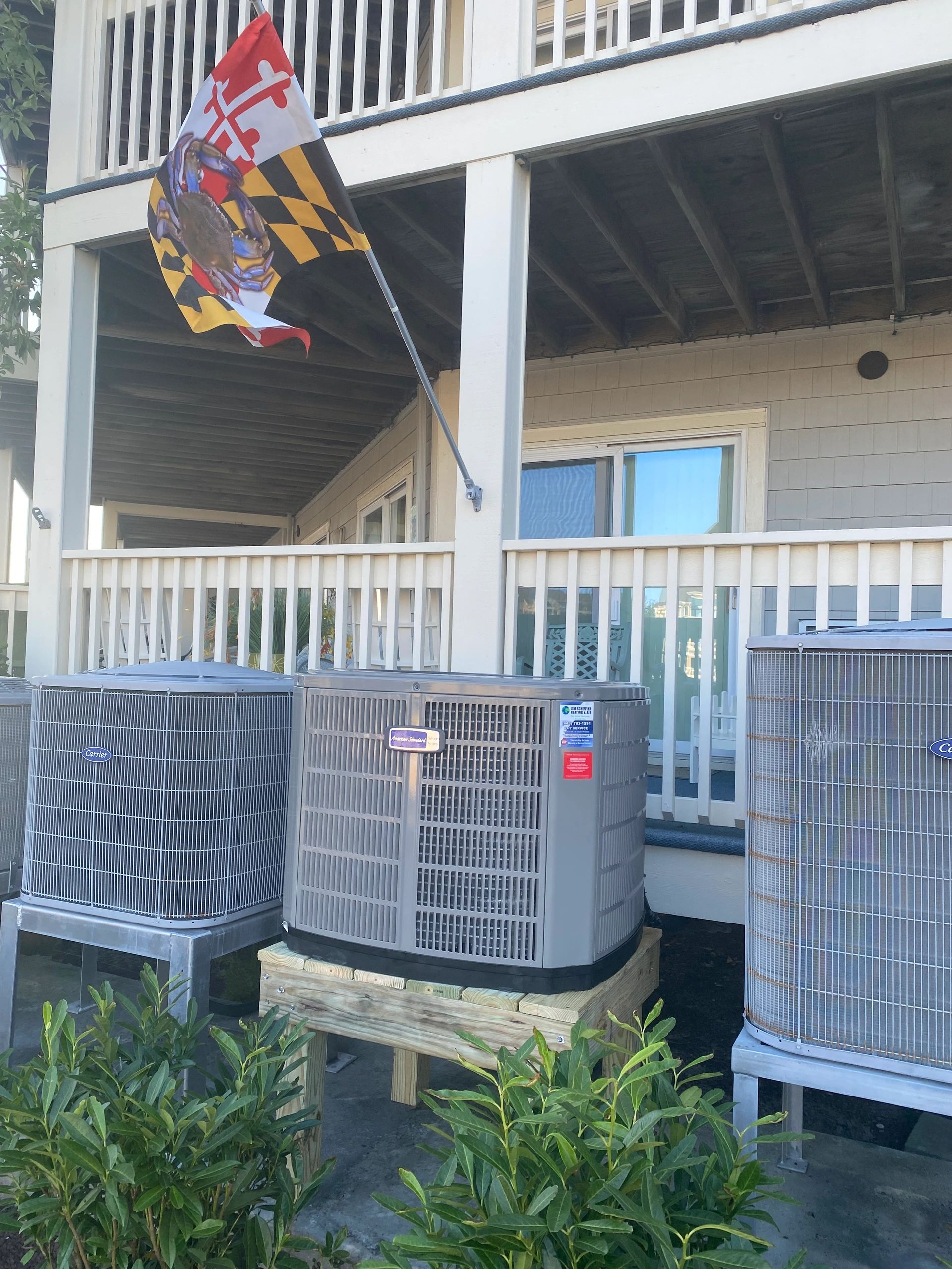 New American Standard heat pump installed under a beach home deck next to existing units, with a Maryland flag flying